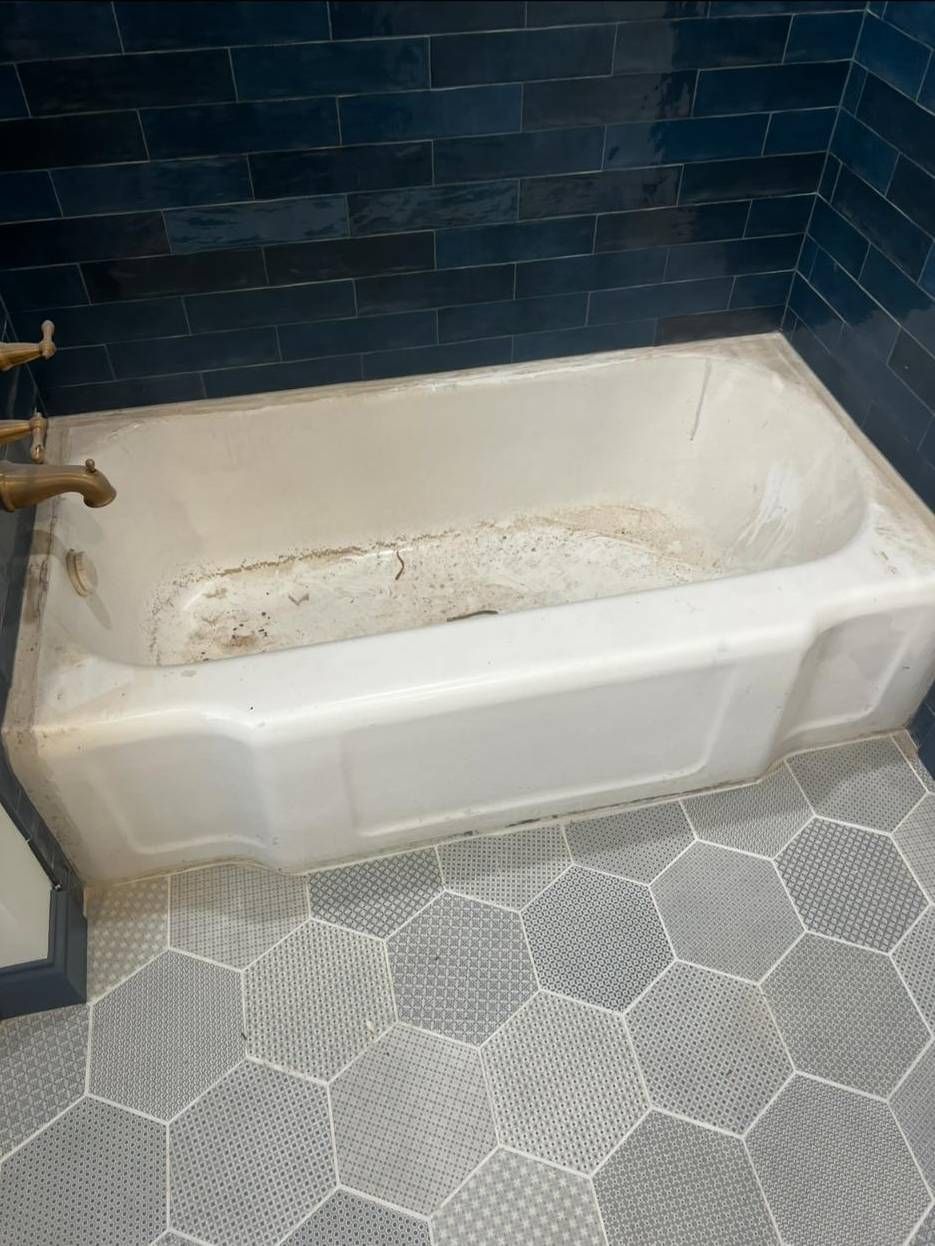A white, stained bathtub with brass fixtures against dark blue subway tiles and a gray hexagonal tiled floor.