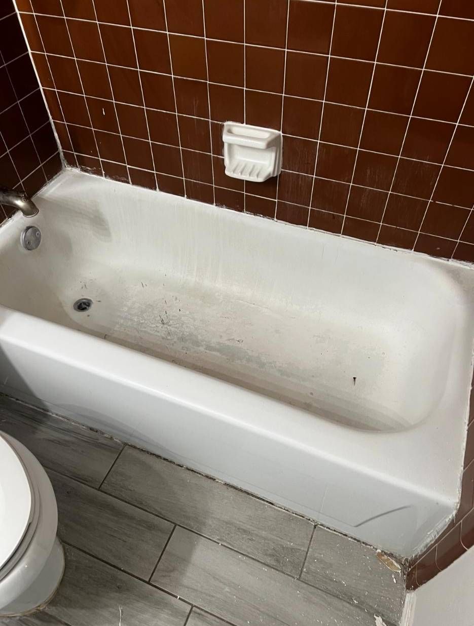 A white bathtub in a bathroom with dark brown tiled walls and grey wood-look floor tiles.