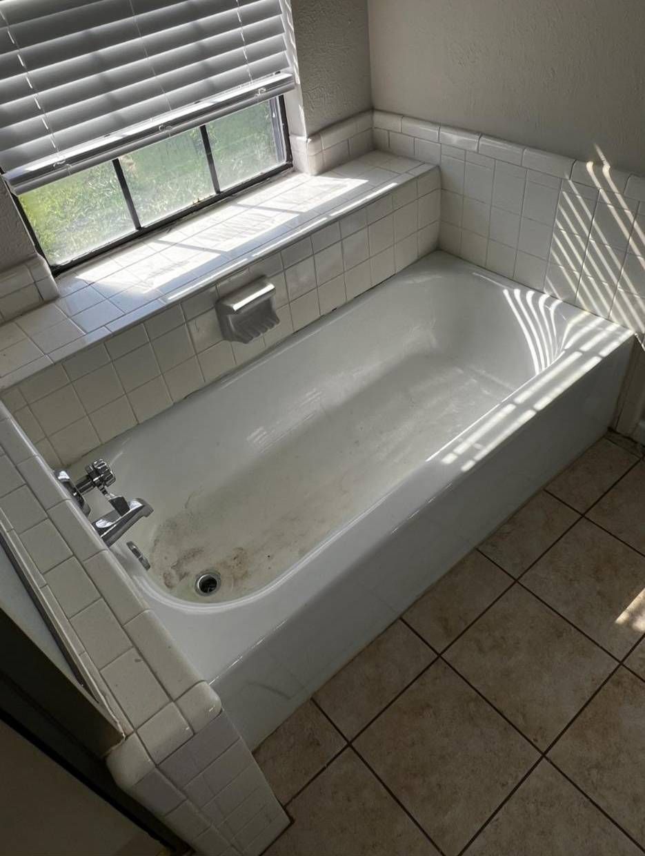 A white built-in bathtub with a tile surround and ledge, positioned next to a window with closed blinds.