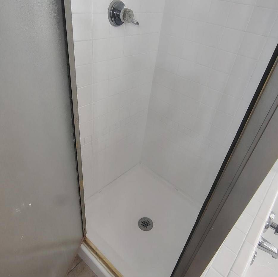 A narrow, white-tiled stall shower featuring a single shower head and a white floor drain.