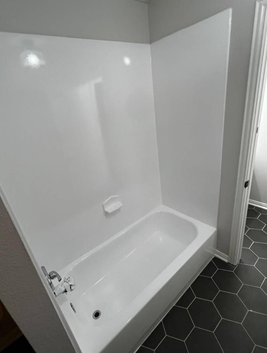 A white bathtub and shower surround set against grey walls with dark hexagonal floor tiles.