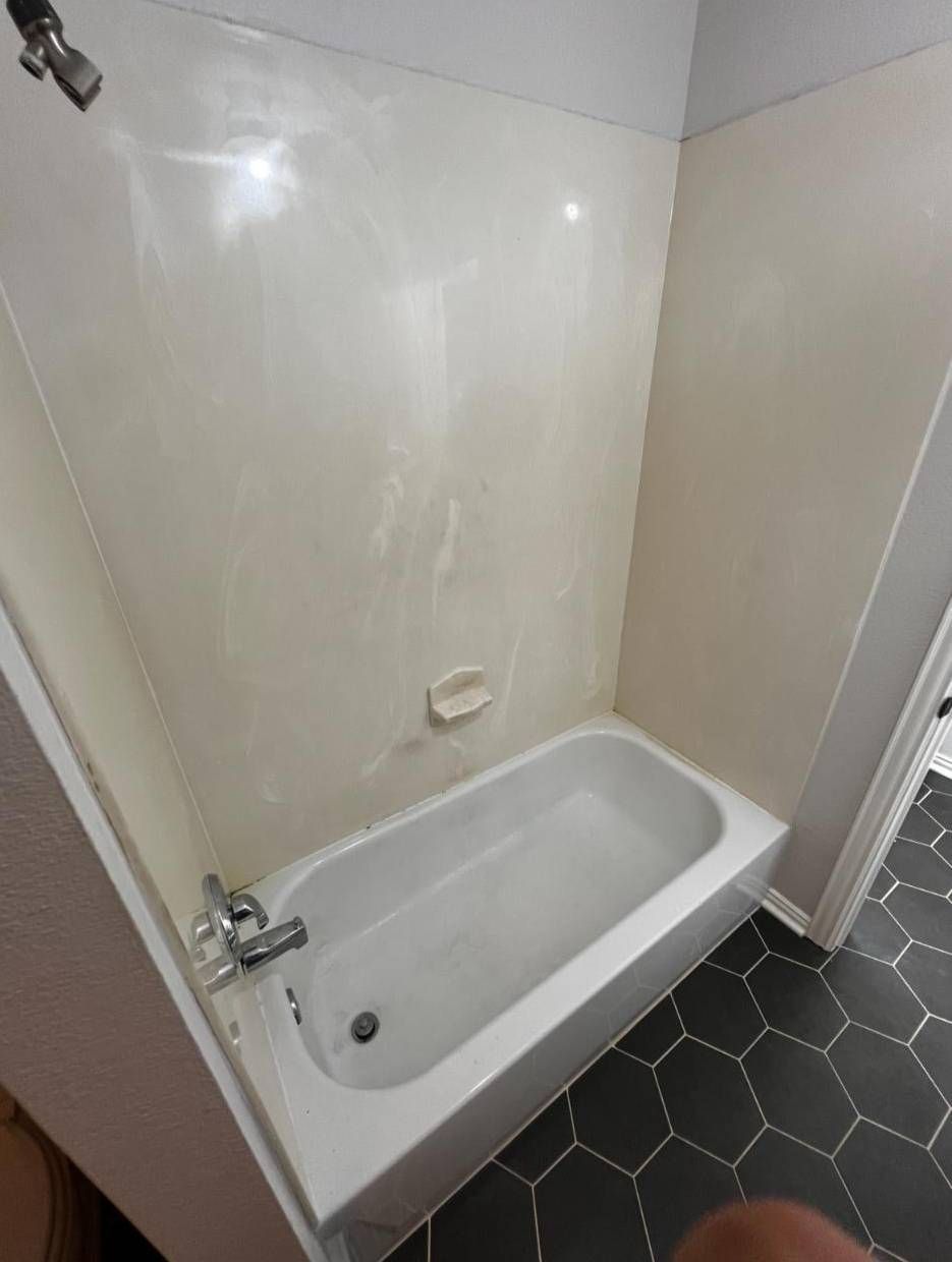 A white bathtub with a wall-mounted shower head and a soap dish, set against cream-colored walls and dark hexagonal tile.