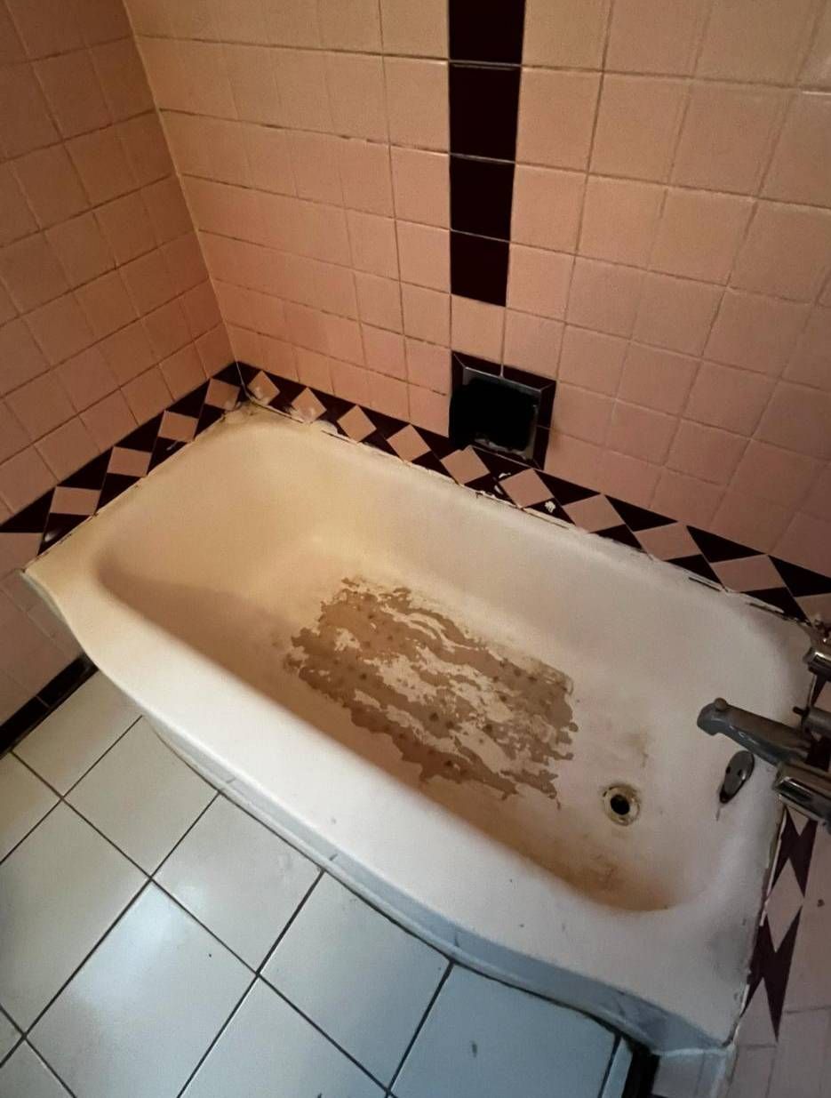A worn, stained white bathtub in a bathroom with pale pink tiled walls and a black-and-white diamond patterned border.