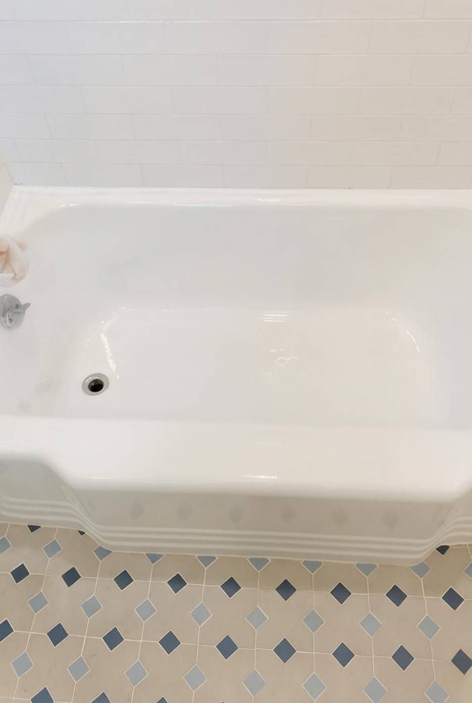 A high-angle view of a white bathtub in a bathroom with white tiled walls and a light-colored floor with blue accents.
