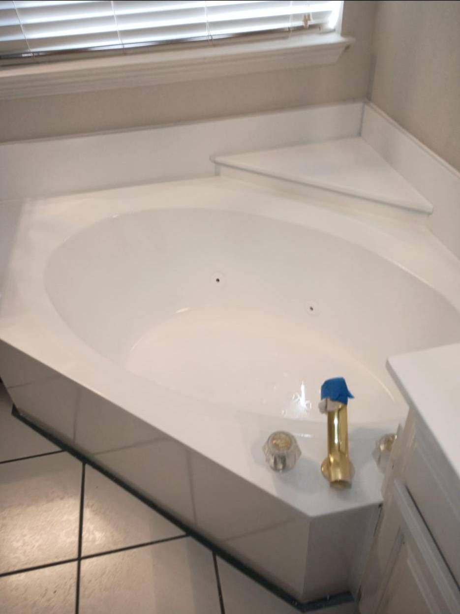 A white corner bathtub with tiled sides and installed faucet hardware below a window with blinds.