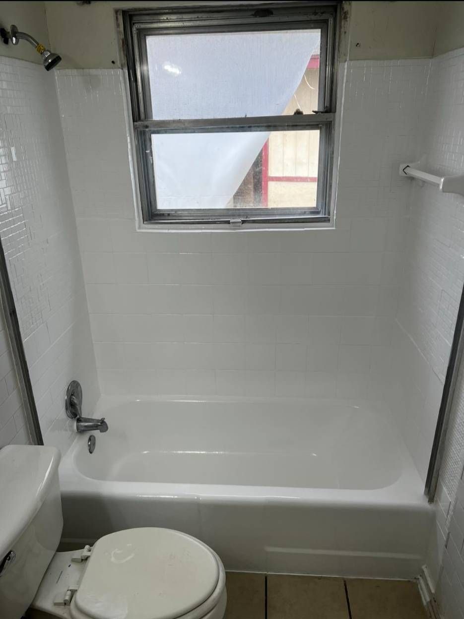 A white bathtub and shower surround with white tiled walls beneath a window, adjacent to a toilet in a bathroom.