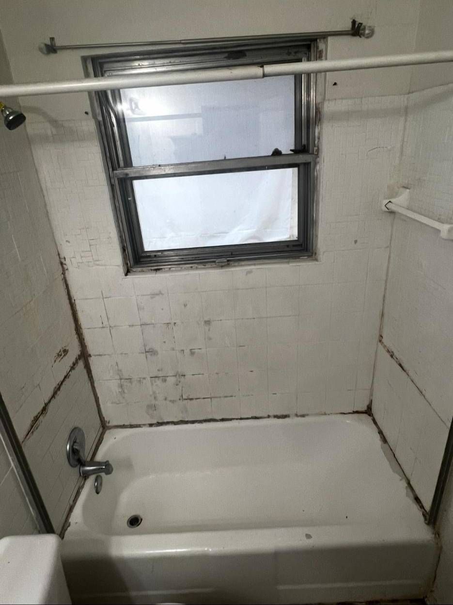 A white bathtub with a silver faucet under a window with a dark frame, surrounded by stained, aging tiled walls.