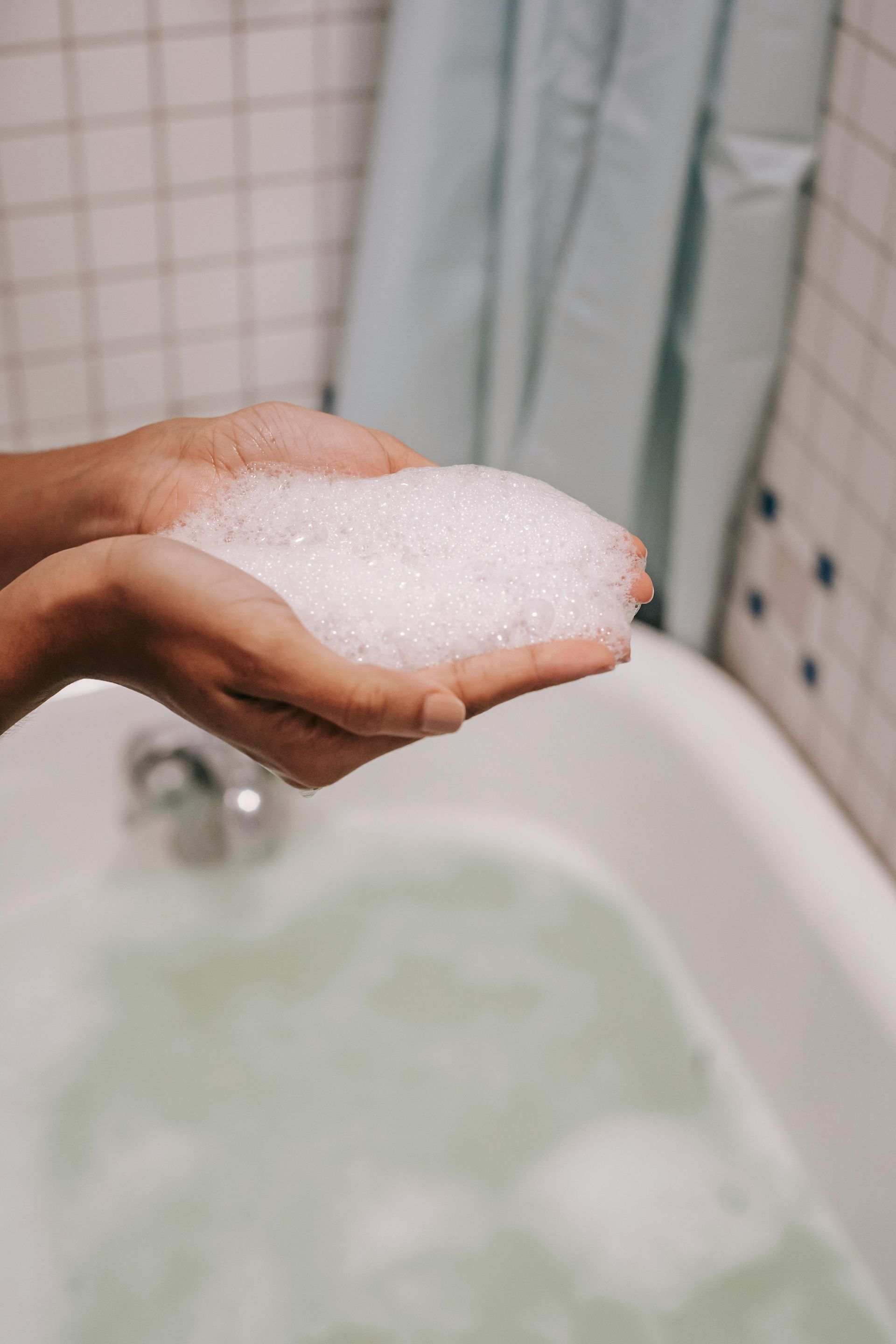 Hands cupped, holding a pile of frothy white bubbles over a bathtub filled with water and foam.