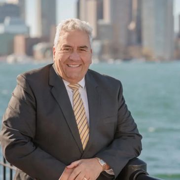 A man in a suit and tie is sitting on a railing with a city skyline in the background.