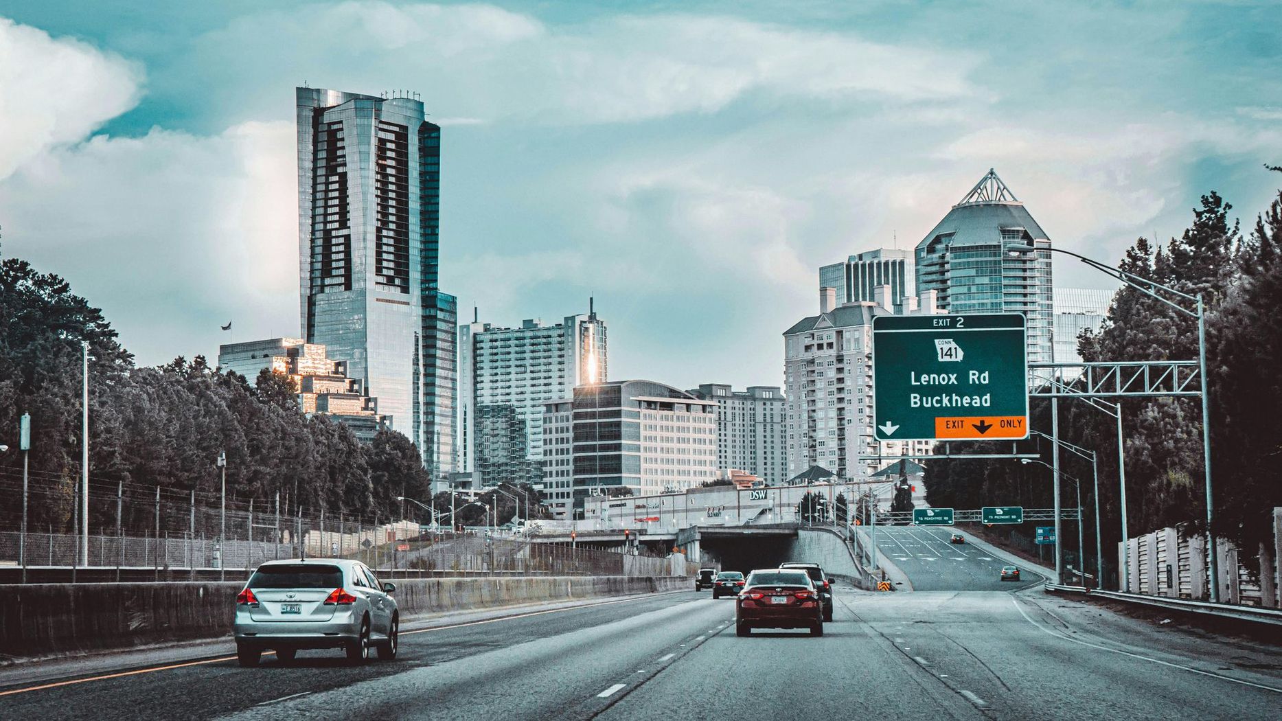 City skyline with highway, cars driving towards buildings under cloudy sky.