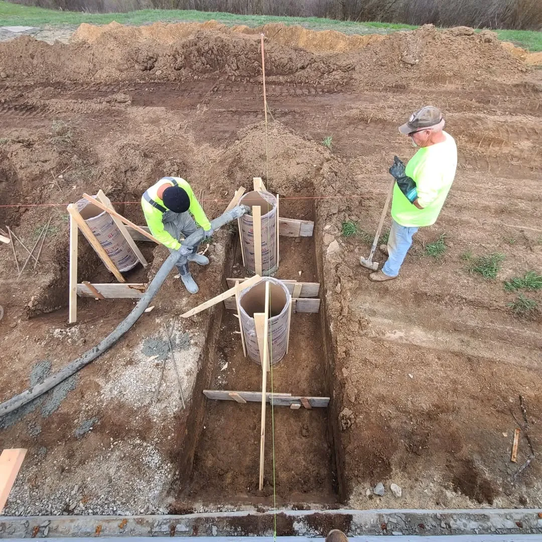Workers pouring concrete into cylindrical footings using a hose and hand tools at a construction site foundation trench.