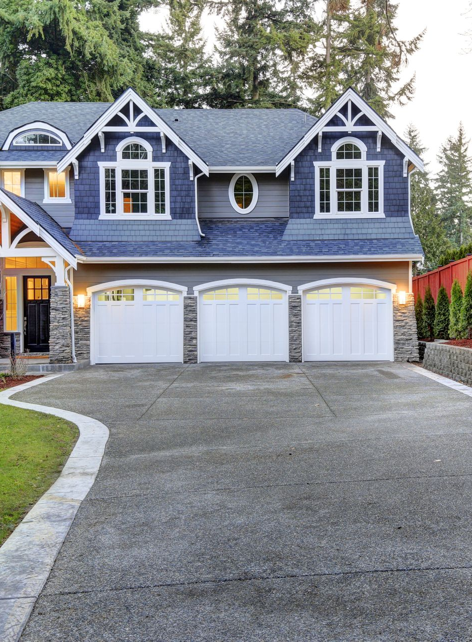 A blue, two-story house with a three-car garage and a stone facade, featuring a paved driveway and a red privacy fence.