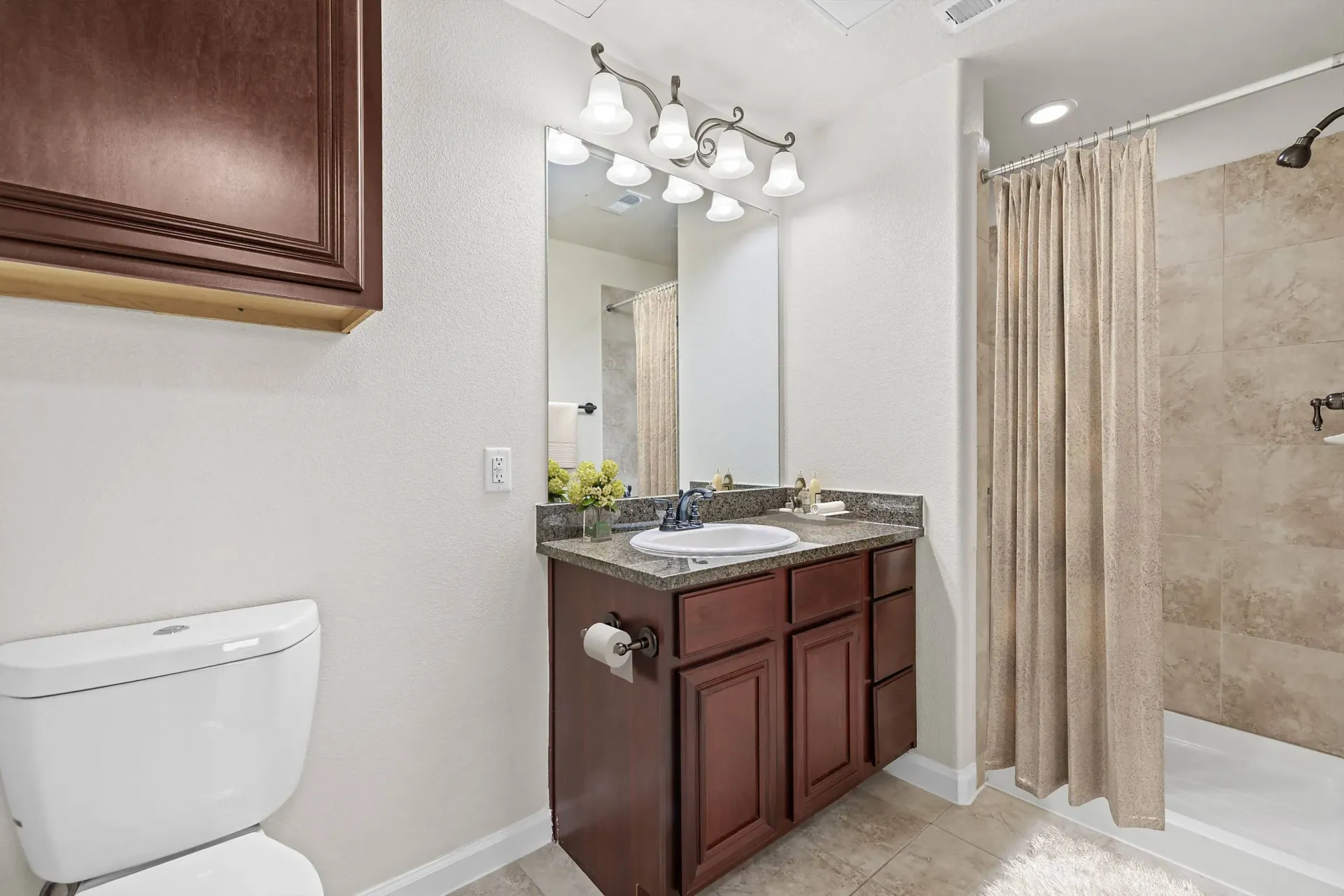 A bathroom featuring dark wood cabinets, a granite countertop, a toilet, and a walk-in shower with a beige curtain.