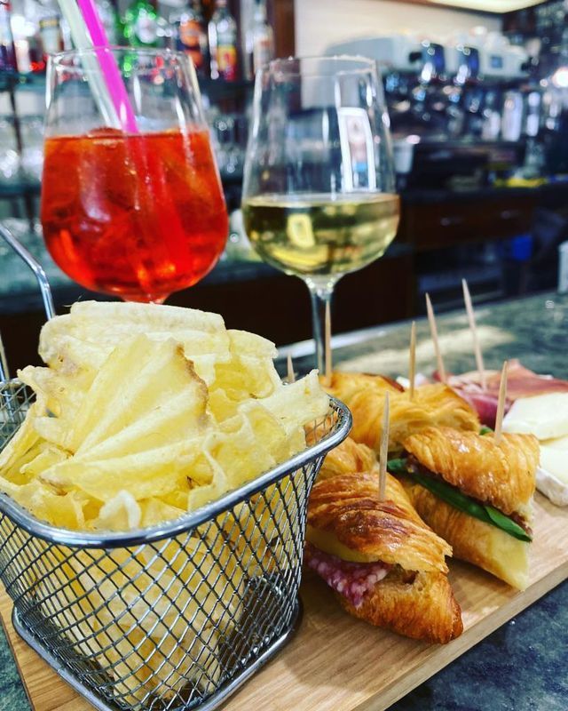 Aperol spritz e vino bianco, con stuzzichini e patatine fritte in un cestino di metallo, su un tagliere di legno in un bar.