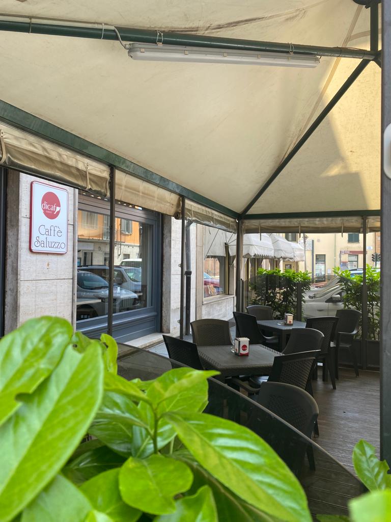 Area salotto all'aperto con tavoli e sedie sotto una tenda da sole, con piante in primo piano.