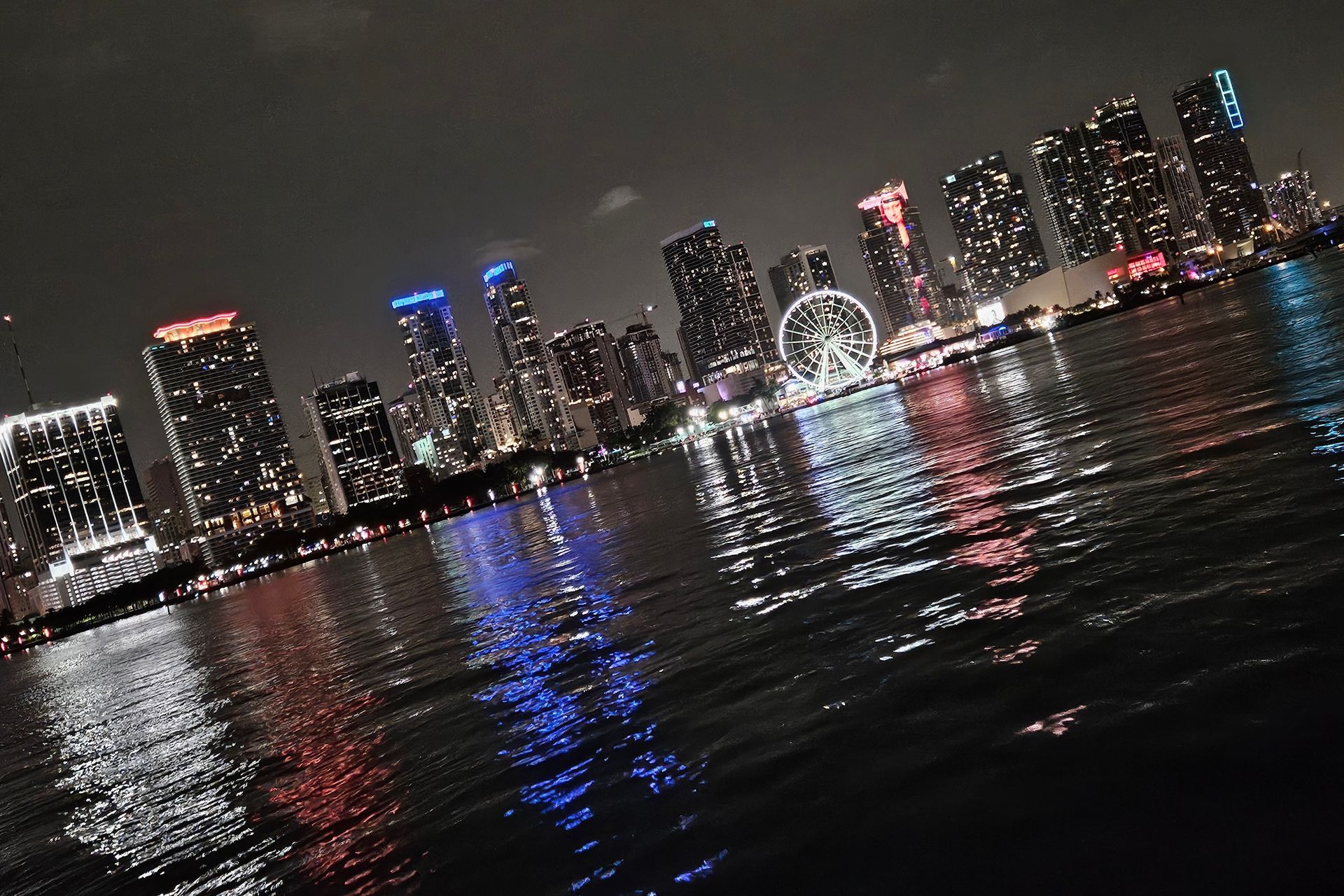 City skyline at night with lights reflecting on water. Ferris wheel visible.
