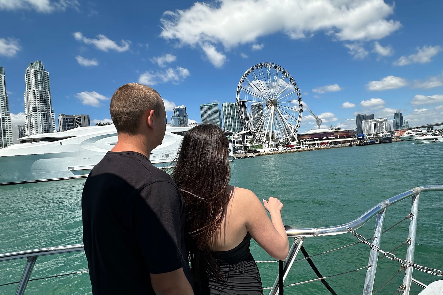 Couple on boat viewing skyline with Ferris wheel under blue sky.
