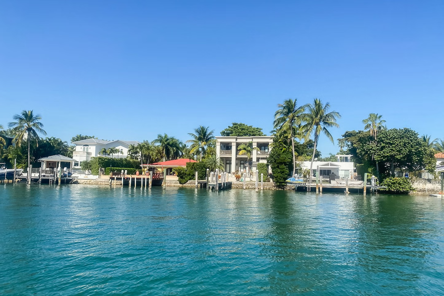 Waterfront houses on a sunny day; turquoise water, docks, and palm trees under a clear blue sky.