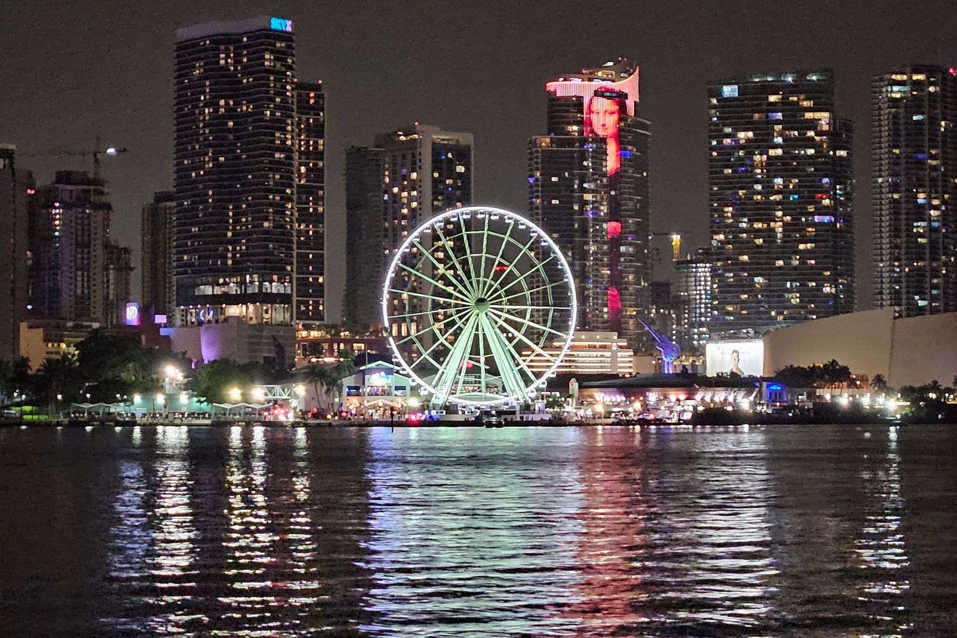City skyline at night, with illuminated Ferris wheel and tall buildings reflected in the water.
