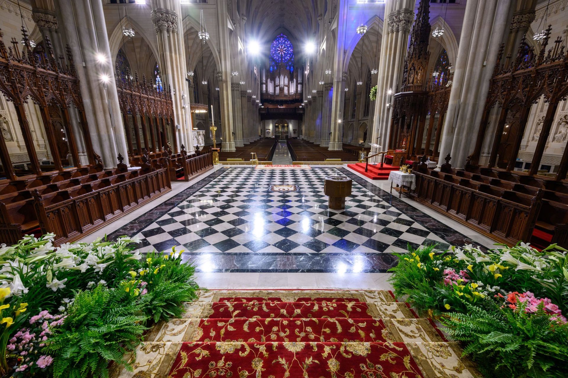 Inside a cathedral, checkered floor leads to the altar, flanked by rows of pews and ornate woodwork.
