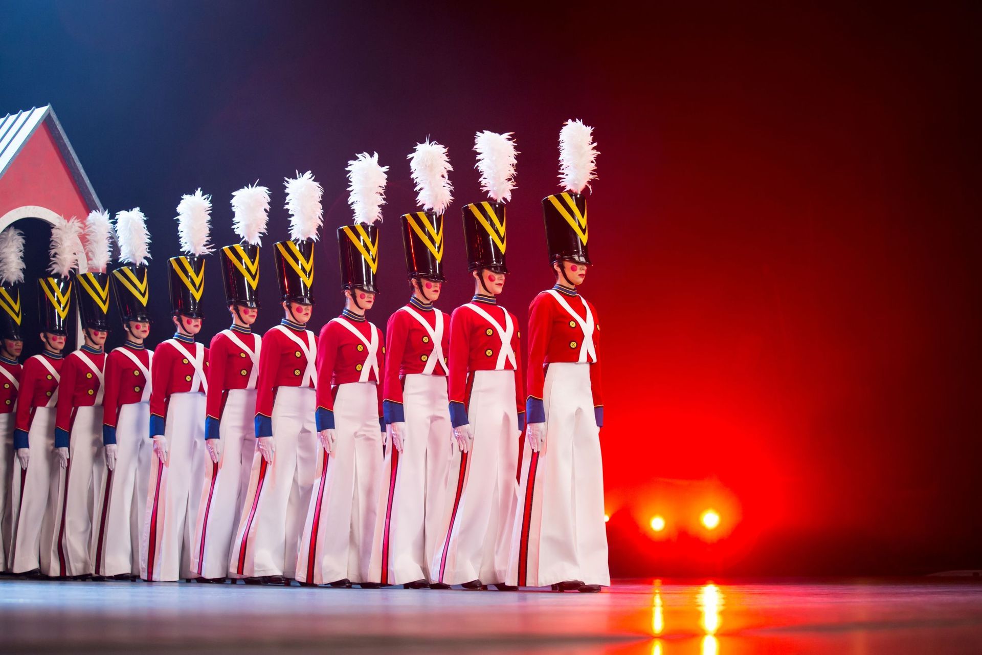 Toy soldier dancers in red coats, white pants, black hats with plumes, marching on stage.