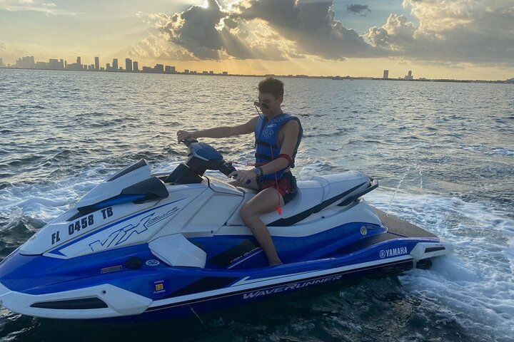 A person on a blue and white jet ski rides on the water with a city skyline in the background.