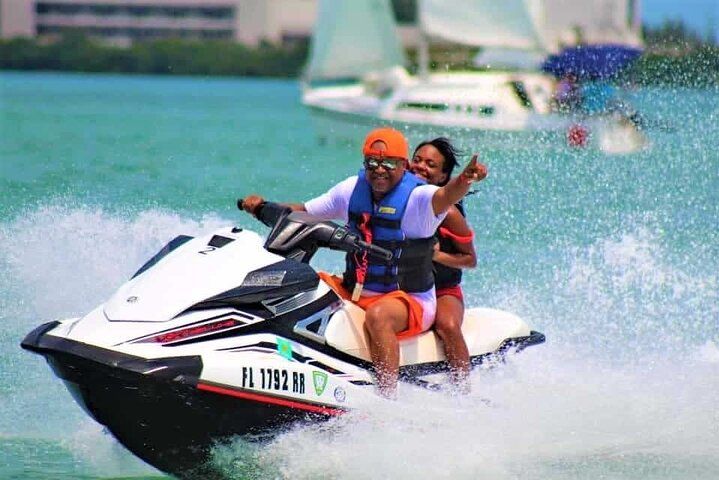 Couple on jet ski, man pointing, woman with arms raised, water splashing, ocean, boat in background.