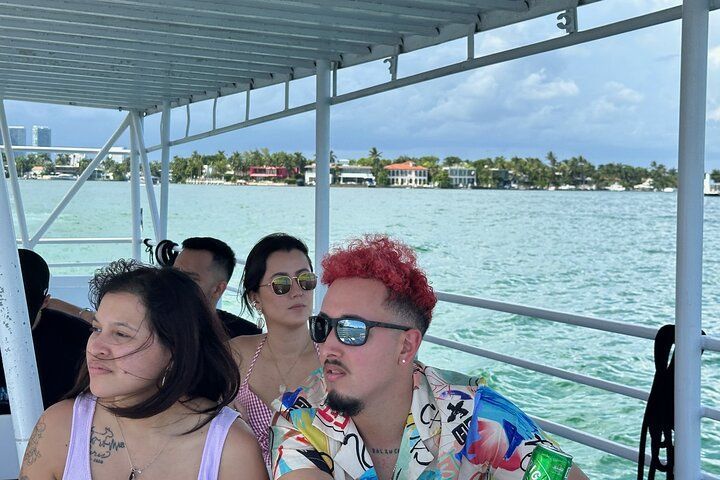 People on a boat looking at the water. One has pink hair and sunglasses. Buildings line the shore.