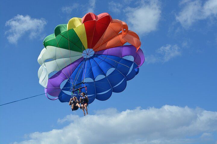 Two people parasailing under a rainbow-colored parachute against a blue sky with clouds.