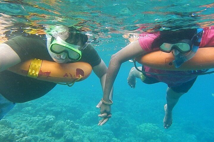 Two people snorkeling, holding hands underwater, near coral reef; one wears a pink shirt.
