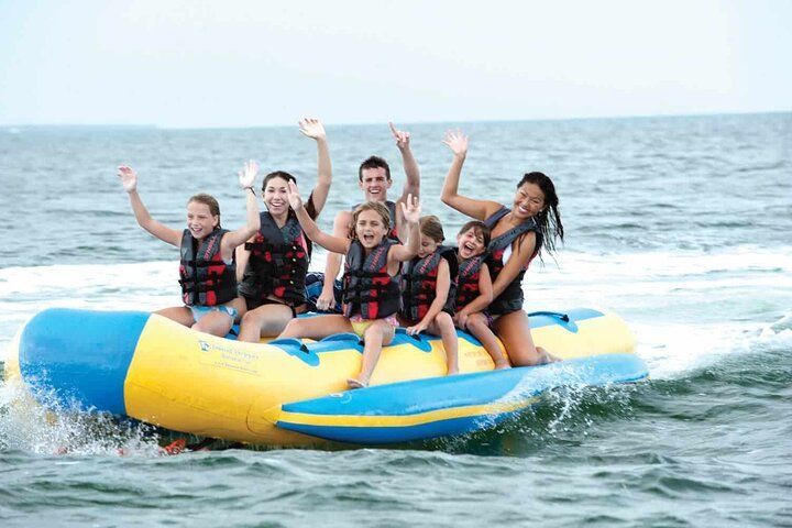 People riding a banana boat on the ocean, waving hands, smiling. The boat is blue and yellow.