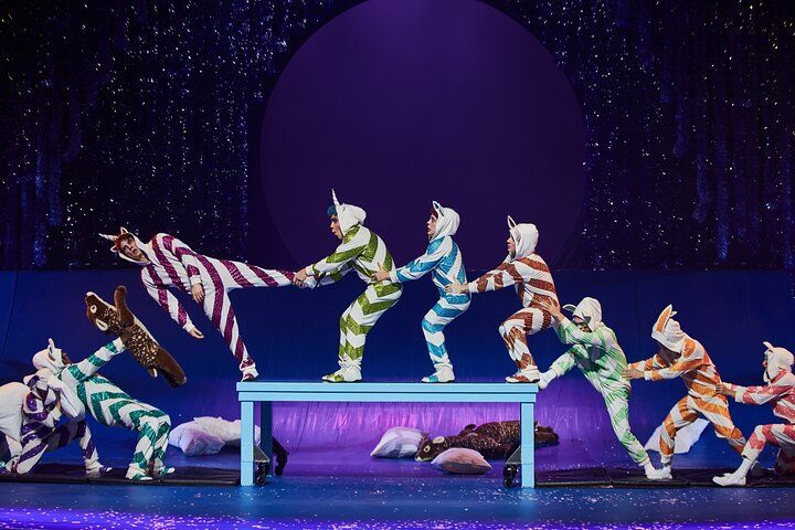 Performers in cat costumes balance on a table, pulling a striped figure under a starry blue backdrop.