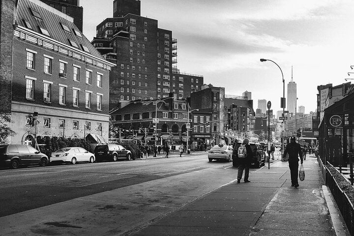 Black and white photo of a city street with buildings, cars, and pedestrians. The Freedom Tower is visible in the background.