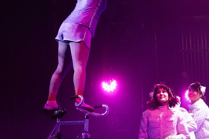 Acrobat stands on a bicycle, balancing, under a spotlight. Two women watch, illuminated in pink.
