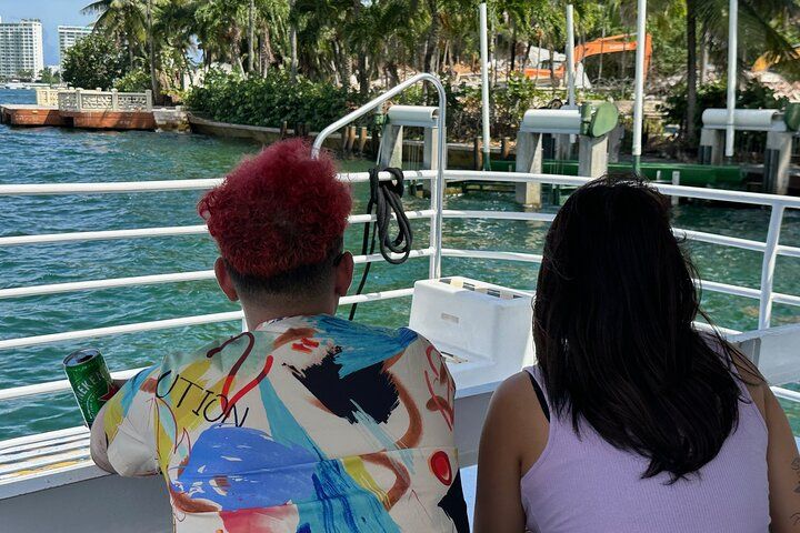 Two people on a boat overlooking water; one with red hair, the other with dark hair. Palm trees and buildings in the background.