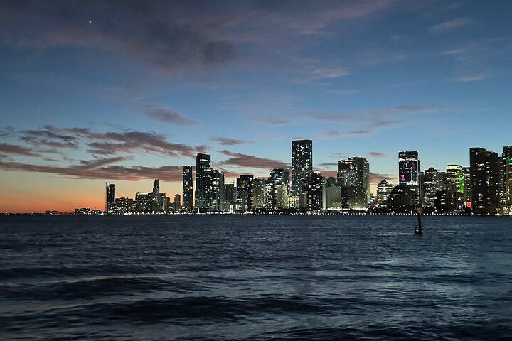 City skyline at dusk, buildings with lit windows against a twilight sky over dark water.