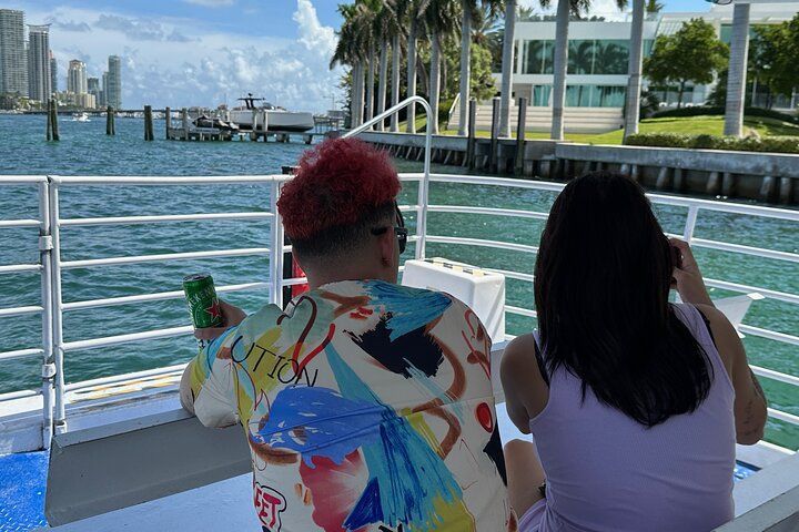 Two people on a boat; man with red hair, colorful shirt; woman in a light purple top, on the water near buildings.