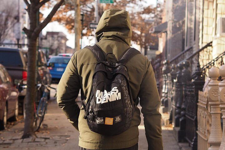 Person walking down a city sidewalk wearing a hooded jacket and backpack; buildings and cars in background.
