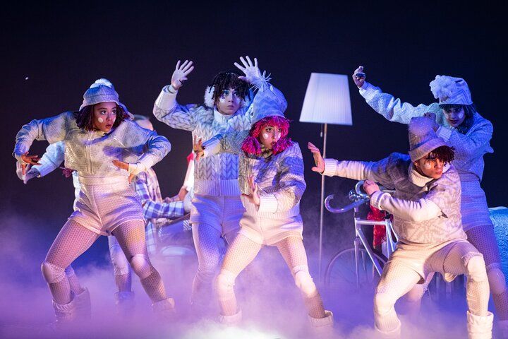 Dancers in white costumes performing on stage with a lamp and bicycle; smoke effect and dramatic lighting.