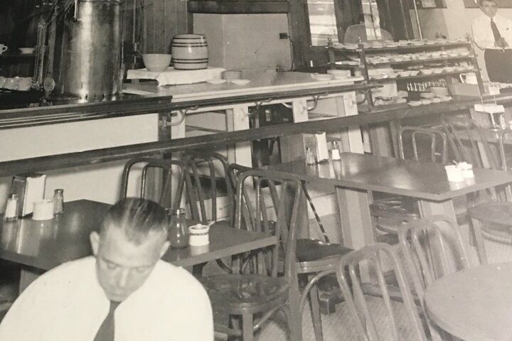 Interior of a diner with tables, chairs, and a man seated, with a counter and shelves in the background. Black and white.