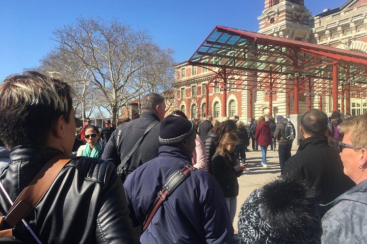 Crowd near a brick building with a glass roof, on a sunny day. People stand, walk, and look around.