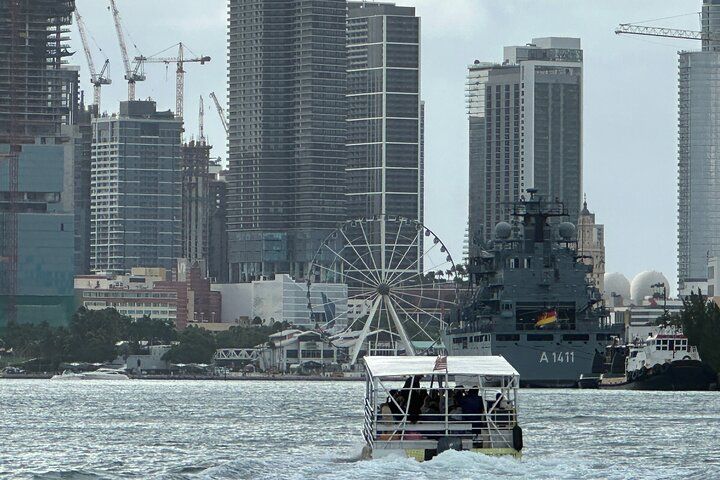 A boat travels on water in front of a city skyline with skyscrapers, a Ferris wheel, and a ship.