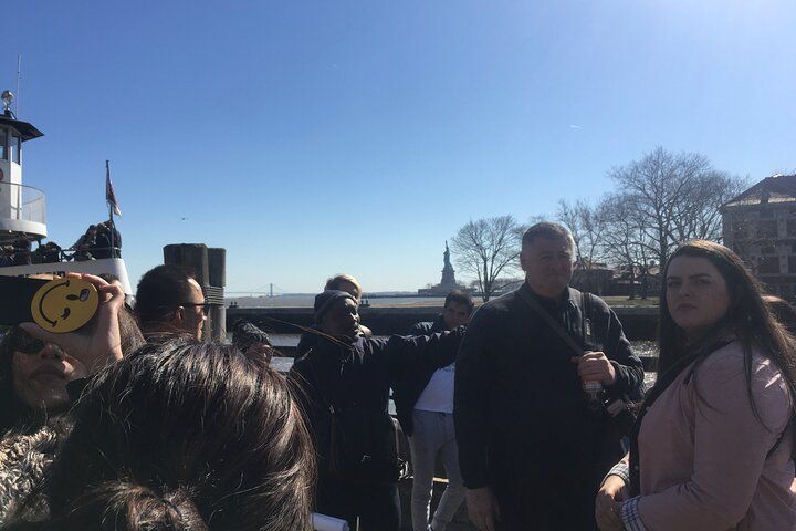People on a dock; Statue of Liberty in the distance. Sunny day; some people pointing towards the monument.