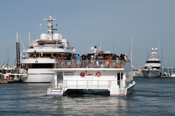 A catamaran boat with passengers sails in front of a large white yacht on a sunny day in a harbor.