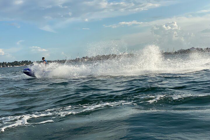 Person on a jet ski speeding across choppy blue water, creating a large spray; coastline in the background.