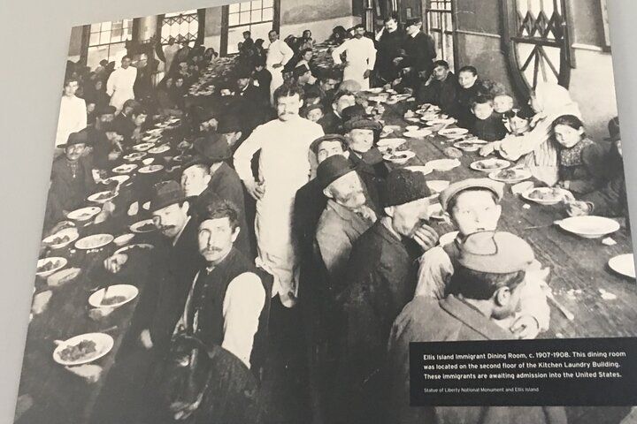 People eating at a long table. Many have plates and some look at the camera. Black and white photo.