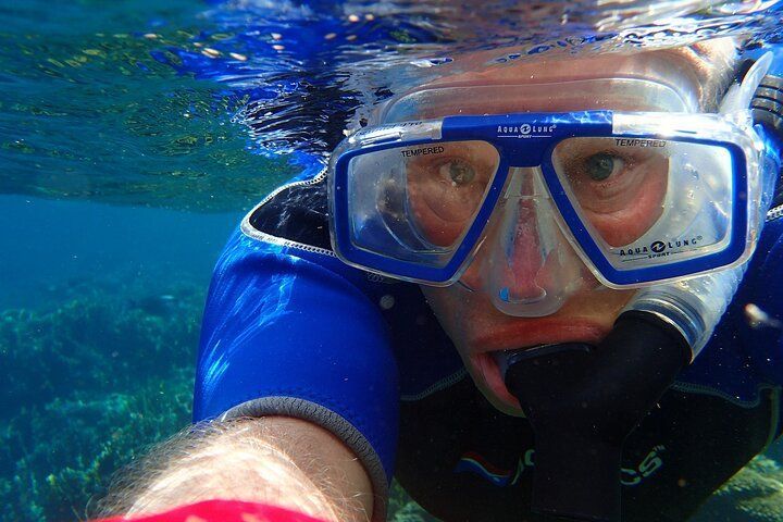 Man snorkeling underwater, wearing blue mask and wetsuit, taking a selfie, with ocean in the background.