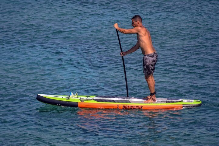 Man paddleboarding on blue water, using a black paddle. The board is orange and green.