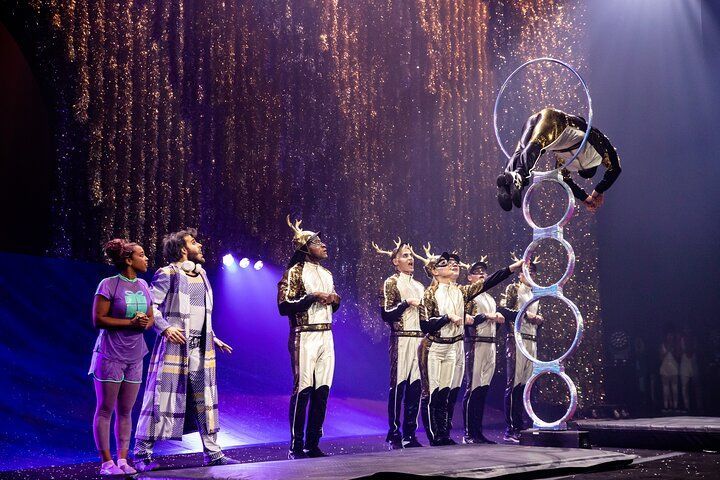 Circus performer in a hoop stack, viewed by a costumed group. Dramatic lighting, sparks in the background.