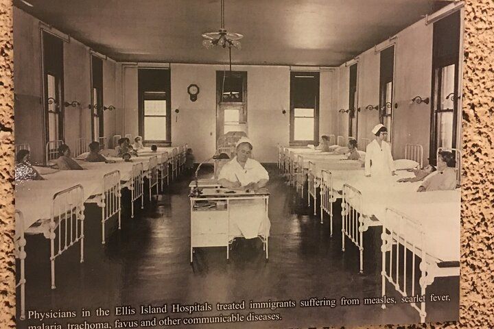 Hospital ward with beds and medical staff. Patients in beds, a nurse sits at a desk, another nurse walks down the aisle.